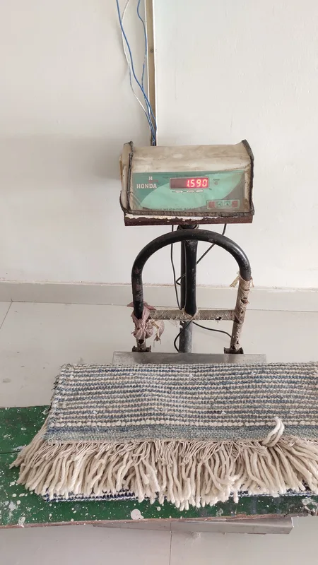 A finished flatweave rug suspended on a hanging scale, digital display reading 1590 grams